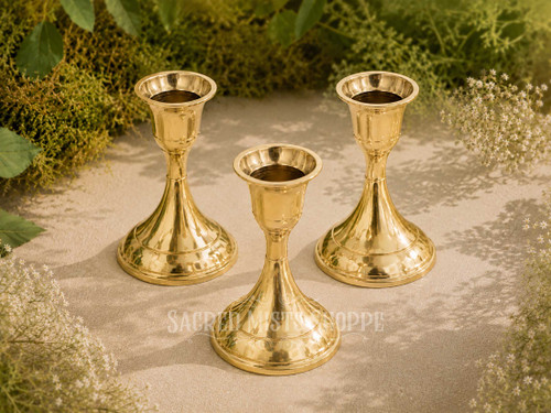 Three polished brass taper candle holders for standard taper candles arranged outdoors on natural stone with greenery and white flowers in bright natural sunlight.