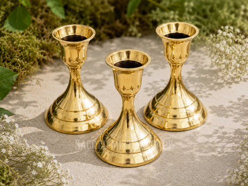Three polished brass taper candle holders with stepped bases for standard taper candles arranged outdoors on natural stone with greenery and white flowers in bright natural sunlight.