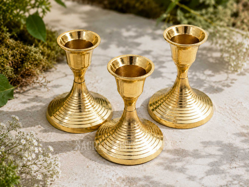 Three polished brass taper candle holders with decorative concentric ring bases for standard taper candles displayed outdoors among greenery and delicate white flowers in natural sunlight.