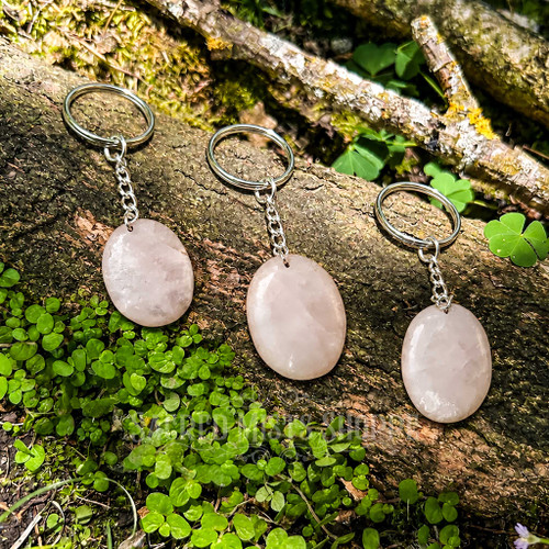 Three natural Rose Quartz worry stone keychains shown from the smooth reverse side on a mossy woodland log, with pale pink polished stones and silver-colored hardware.