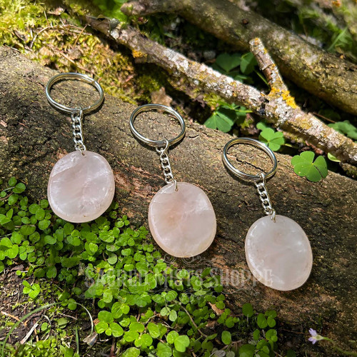 Three natural Rose Quartz worry stone keychains displayed on a mossy woodland log, showing polished pale pink crystals with thumb indentations and silver-colored keychain hardware.