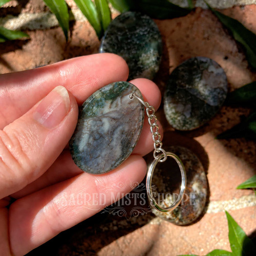 Close-up of a natural Moss Agate worry stone keychain held in a hand, showing its polished oval shape, green and gray moss-like patterning, and silver-colored keychain hardware.
