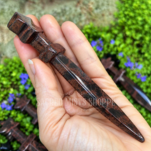 Mahogany Obsidian crystal athame held in hand to show its 5.25 inch size, polished pointed blade, crossguard, and dark patterned stone.