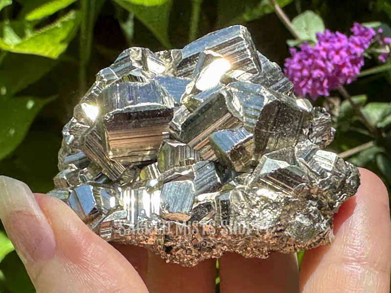 Ethically sourced cubic Peruvian pyrite cluster held between fingers, sparkling in the sunlight with lush greenery in the background. Known for attracting abundance and shielding negative energy.