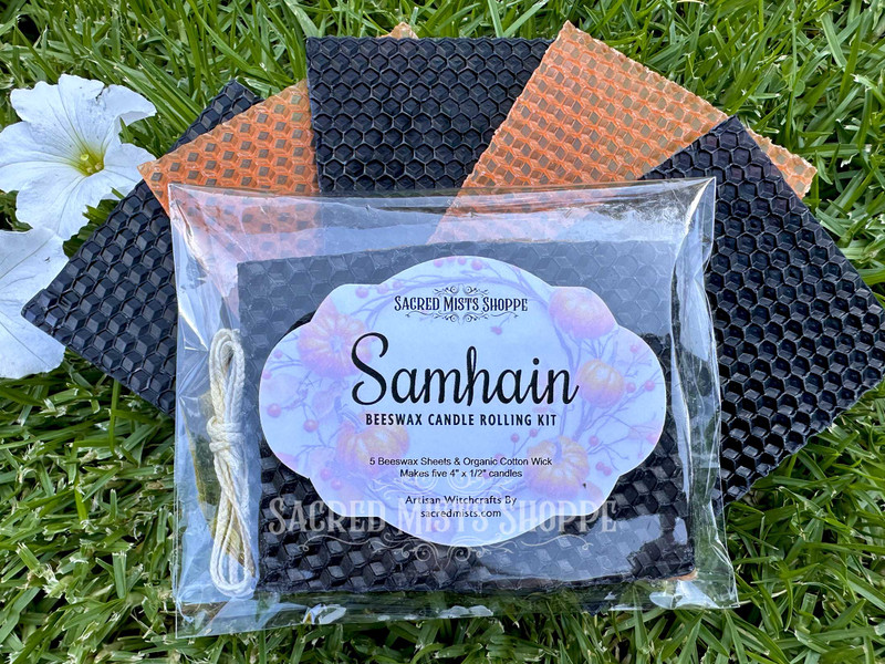 a close up of one samhain and halloween beeswax candle rolling kit lying atop 5 sheets of beeswax fanned lying on grass with white flowers nearby. perfect for making your own samhain, halloween, or ancestor spell and ritual candles.