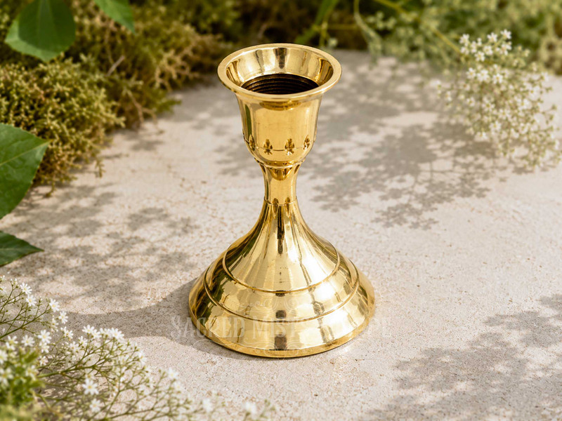 Polished brass taper candle holder with stepped base for standard taper candles displayed outdoors on natural stone surrounded by greenery and delicate white flowers in warm sunlight.