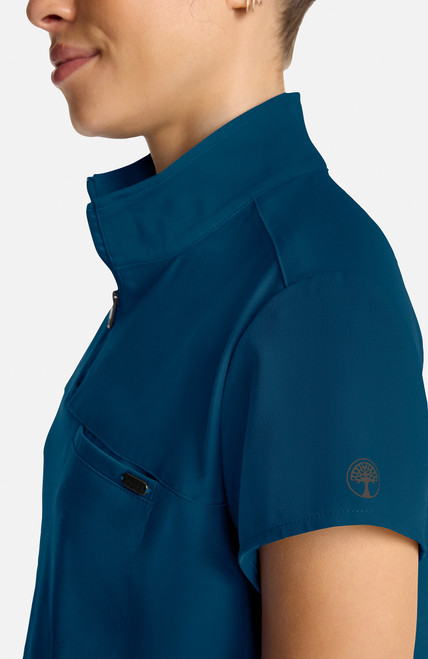 The HH752 Quest Bella Women’s Partial Zip Stand Collar Top is shown in a close‑up view emphasizing its Caribbean Blue short‑sleeve construction, smooth partial‑zip collar, and practical chest pocket holding a pen. The clean, professional detailing reinforces its purpose as a dependable uniform top for healthcare, hospitality, and other active roles.
