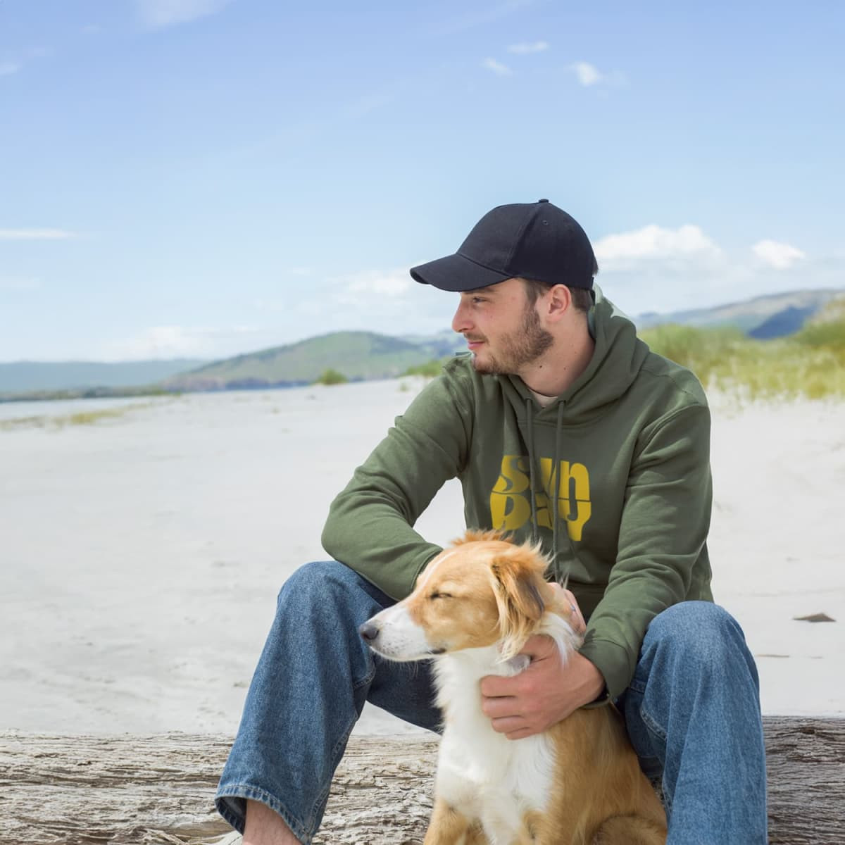 A person wearing an olive green unisex hoodie sits on a beach with a dog, surrounded by sand and hills in the background.