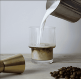 A glass tumbler with a ribbed design filled with coffee and milk, accompanied by coffee beans and a pouring jug.
