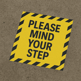 A yellow floor decal sticker with black striped edges and text instructing to "MIND YOUR STEP."