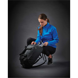 A woman in a bright blue softshell jacket is opening a black duffel bag. She is crouched down, focused on the bag.