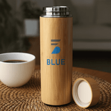 A drink bottle in bamboo finish with a silver lid and a logo. It sits on a woven surface near a cup.