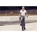 A woman in a white polo shirt rides a bike in a skate park, wearing grey pants and a sweater tied around her waist.