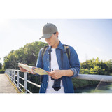 A man wearing an olive green cap and denim shirt examines a map while standing on a bridge with a camera strap.