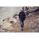 A woman walks along a rocky path by the water, wearing a black rain jacket and casual jeans.