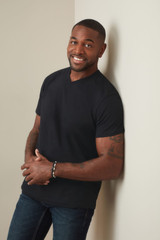 A man wearing a black classic V-neck T-shirt, standing against a wall with a friendly smile.