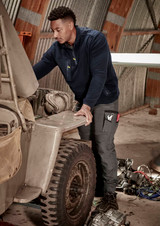 A man in a navy 1/4 zip polar fleece works on an engine in a workshop, wearing work pants with pockets.