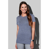 A woman wearing a blue recycled sports T-shirt, smiling and posing against a grey background. The shirt features a logo.