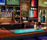 A bar mat featuring a tropical design in vibrant colours, laid out on a bar with drinks nearby.