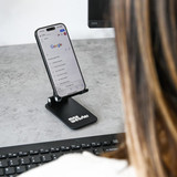 A black folding desktop mobile phone stand holding a smartphone, positioned on a desk near a computer keyboard.