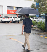 A person walking under a large black golf umbrella, with cars and a post office in the background.