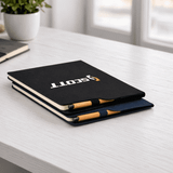 A black and navy A4 notebook with a logo, stacked on a desk next to a small pot plant.