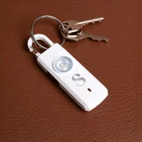 A white safety alarm keychain with a circular light, attached to a set of silver keys on a brown leather surface.