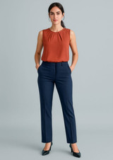 A woman in an orange sleeveless top and navy tapered leg adjustable waist pants, standing with hands in pockets.