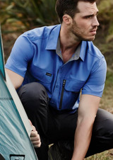 A short-sleeve, blue button-up shirt with one chest pocket and logo, worn by a man outdoors near a tent. A short-sleeve, blue button-up shirt with one chest pocket and logo, worn by a man outdoors near a tent.