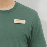 A wooden badge is attached to a green shirt, featuring a branding logo and a name.