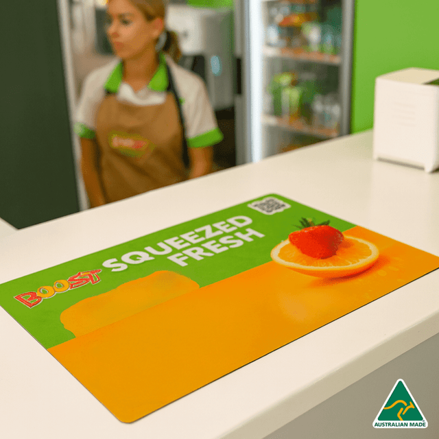 A vibrant orange counter mat featuring fruit graphics and a logo, set on a retail counter. A vibrant orange counter mat featuring fruit graphics and a logo, set on a retail counter.