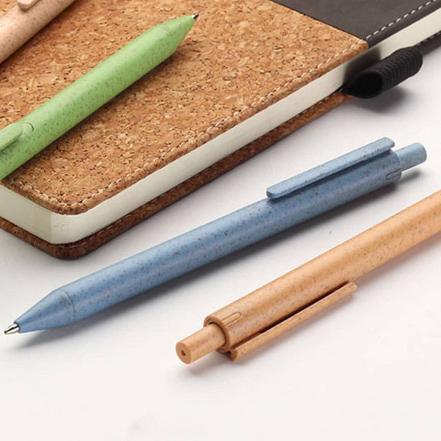 A collection of eco-friendly pens in green, blue, and tan, placed beside a cork notebook. The pens have a logo.