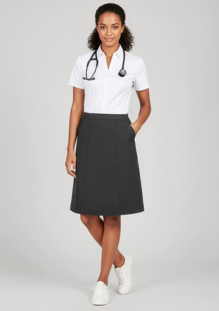 A woman in a white shirt and grey cargo skirt stands smiling, wearing white shoes and a stethoscope around her neck.