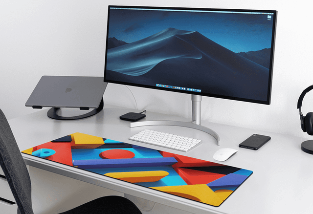 A vibrant desk mat featuring colourful geometric patterns sits on a sleek, modern desk with a computer monitor and accessories.