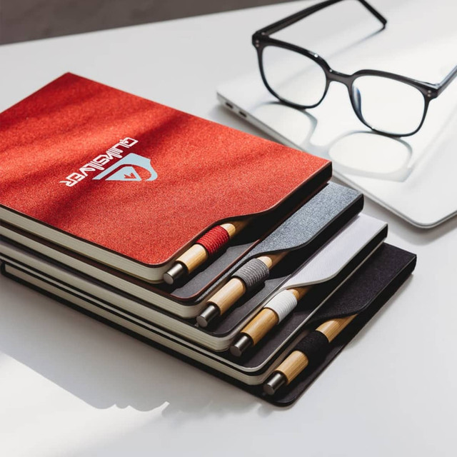 A set of A5 notebooks in red, black, white, and grey with matching pens, placed next to glasses and a laptop.