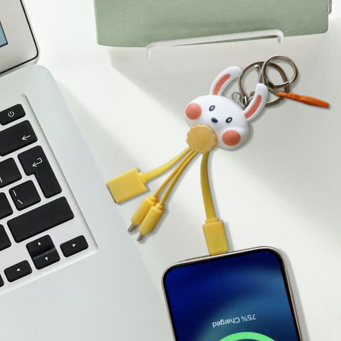 A custom-shaped PVC charging cable in yellow with a bunny keyring, resting on a desk next to a laptop and smartphone.