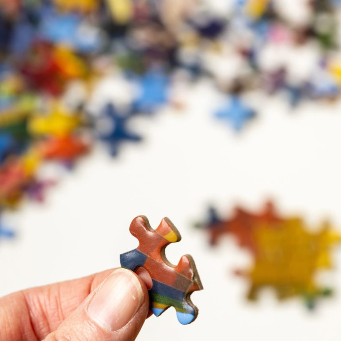 A hand holding a colourful jigsaw puzzle piece, with many more pieces scattered in the background.