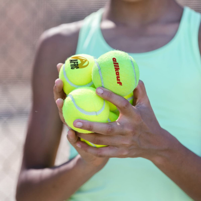 A person holding four bright yellow tennis balls, featuring different logos, against a sports background.