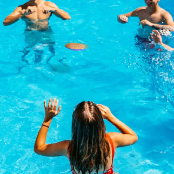 A water bouncing disc in mid-air above a pool, with people in swimwear enjoying the water around it.