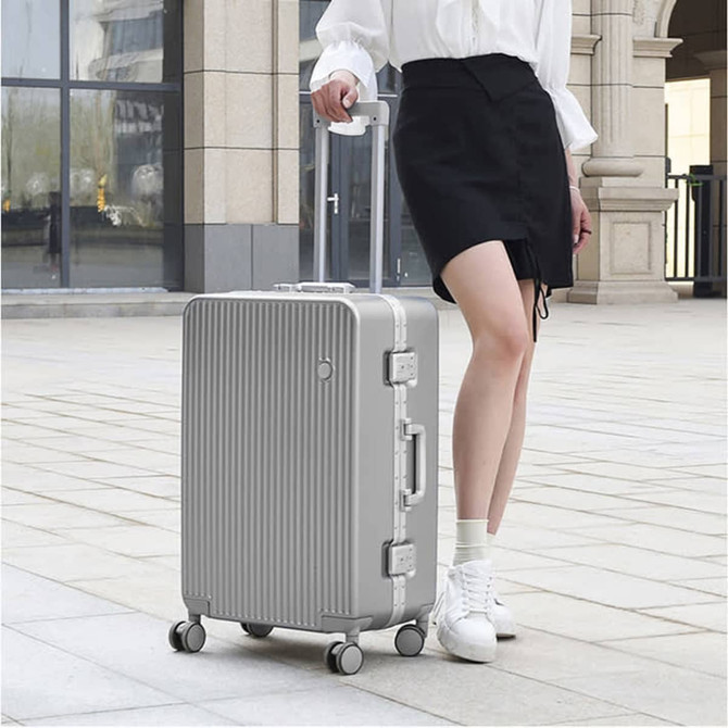 A silver ribbed ABS suitcase with wheels stands on a tiled surface next to a person wearing a skirt and sneakers.