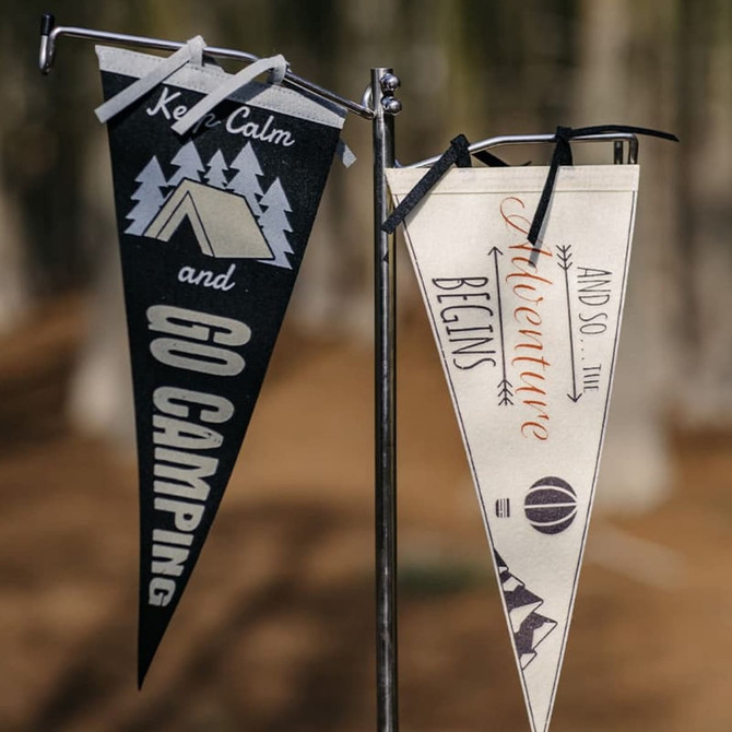Two felt pennants displayed on a stand: one black with a camping theme and one cream with adventure-themed graphics.