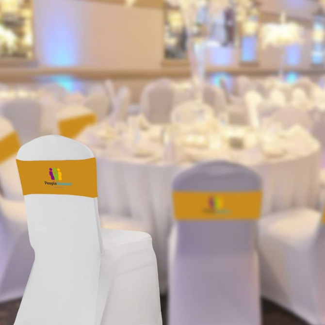 A white chair cover with a yellow band featuring a logo, set against a blurred background of an elegant event space.