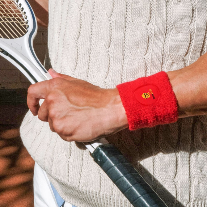 A red Terry Sport Wristband is worn on a wrist, alongside a tennis racket, against a textured background.