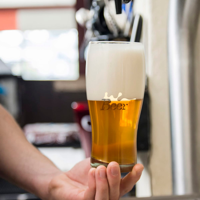 A drink glass filled with beer, featuring a frothy white head and a branded logo, set against a bar background.