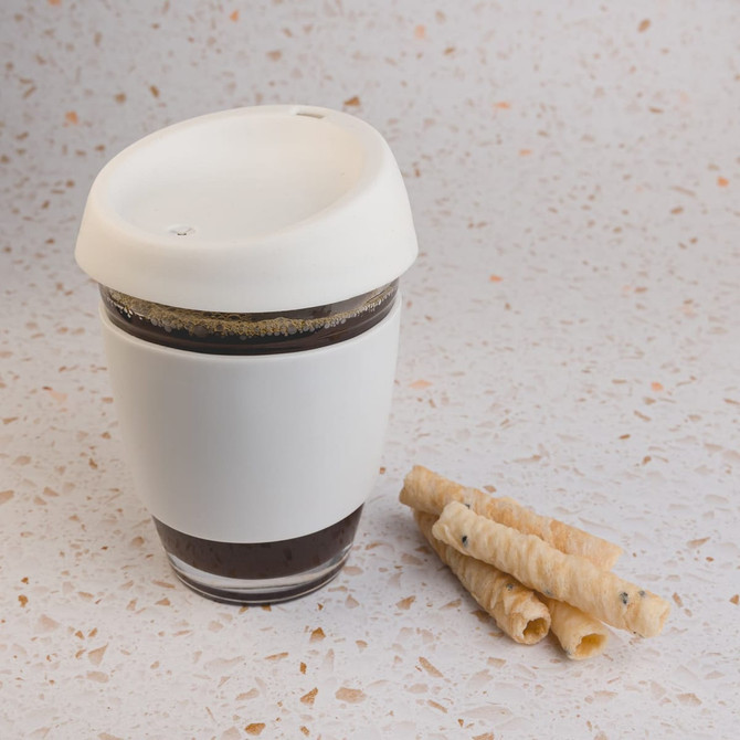 A reusable coffee cup in white with a silicone lid, alongside rolled wafer snacks on a speckled surface.