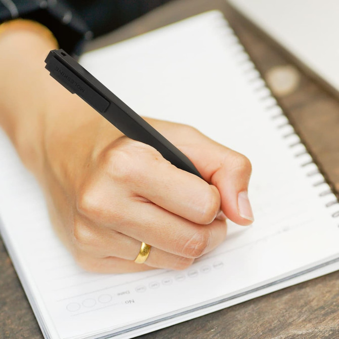 A black Moleskine Go Pen held in a hand, writing on a spiral-bound notepad. The background features a wooden surface.