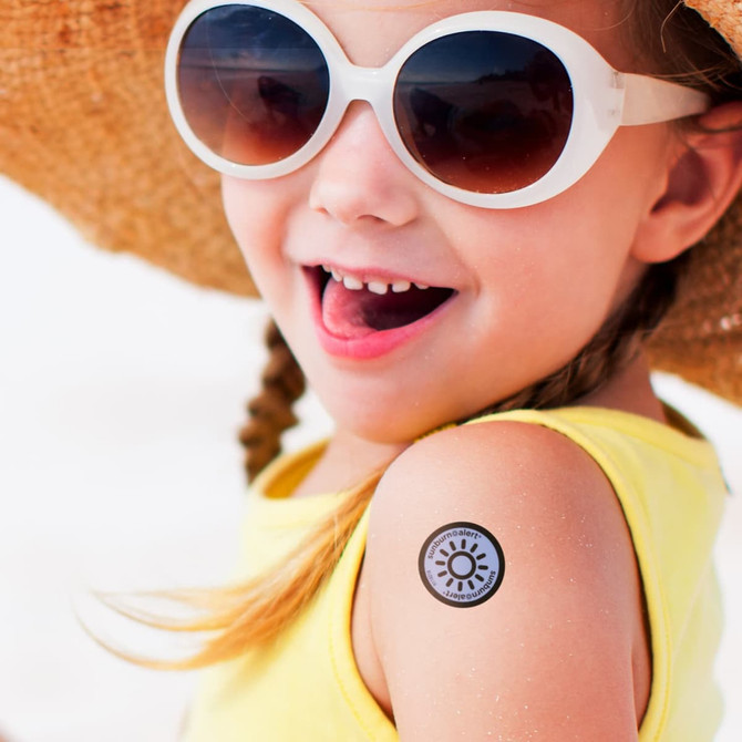 A girl in sunglasses smiles with a sunburn alert sticker on her bare shoulder, wearing a yellow top and a straw hat.