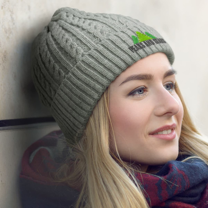 A light green knit beanie worn by a woman, featuring a logo on the front, with a scarf around her neck.