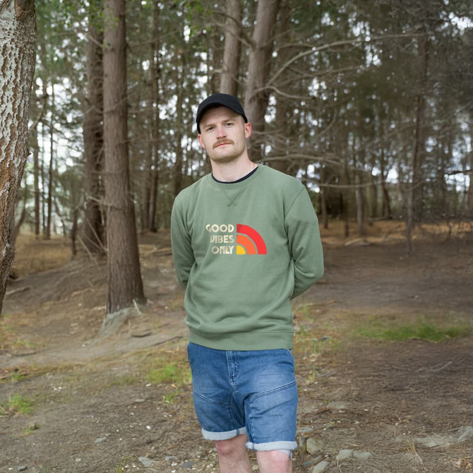 A man stands in a forest wearing a green unisex sweatshirt with a logo. He pairs it with denim shorts.