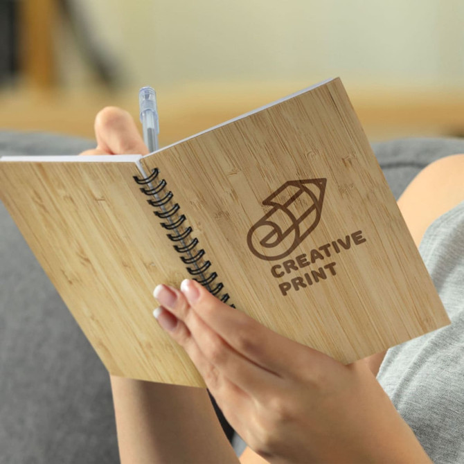 A medium bamboo notebook with a spiral binding, held by someone writing with a pen. It features a printed logo.